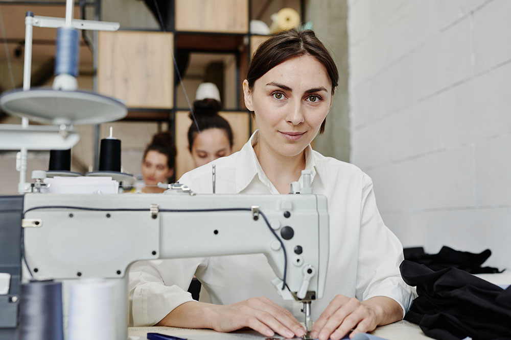 Young contemporary seamstress looking at you while sitting by workplace with electric sewing machine and making or repairing clothes